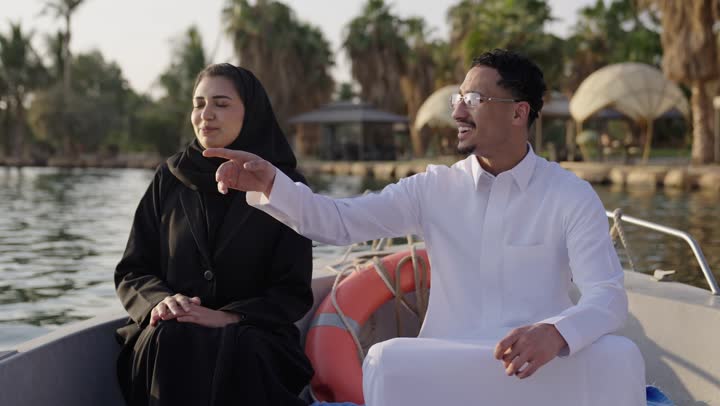 الاشارة الى شيء ما، قضاء وقت ممتع فى الخارج، حوارات ونقاشات في الأجواء الطبيعية،ايماءات السعادة و السرور، رجل عربي خليجي سعودي يرتدي الثوب الابيض يجلس فى الهواء الطلق على مركب برفقة زوجته