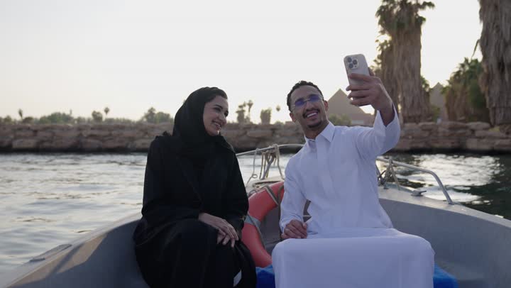 Documenting travel moments and commemorative photography, a tourist Nile trip, an Arab Gulf couple from Saudi Arabia wearing traditional attire sitting inside a riverboat taking pictures with a mobile phone, a calm recreational atmosphere, domestic tourism and river stroll.