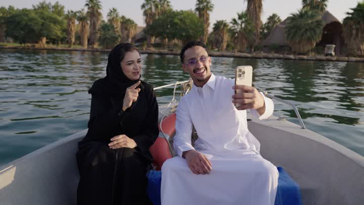 رحلة نيلية سياحية، توثيق لحظات السفر والتصوير التذكاري، زوجان عربيان خليجيان سعوديان يرتديان الزي التقليدي يجلسان داخل مركب نهري يلتقطان الصورعبر الهاتف المحمول، أجواء ترفيهية هادئة، سياحة داخلية وتنزه نهري