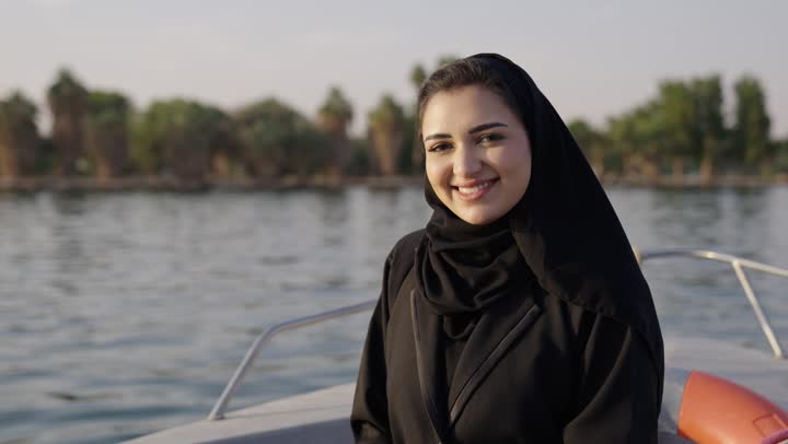 Looking at the camera with gestures of happiness and joy, having a good time outdoors, an Arab Gulf Saudi woman wearing an abaya and hijab sitting inside a boat on a Nile trip, weekend holiday.