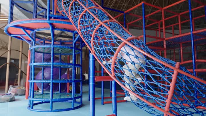 An amusement park that is safe and equipped for children's safety, a young Arab Saudi Gulf boy dressed in casual attire is climbing inside a cylindrical tunnel made of colorful ropes, preparing to start a new adventure in the fun games, enjoying playing in the amusement park.