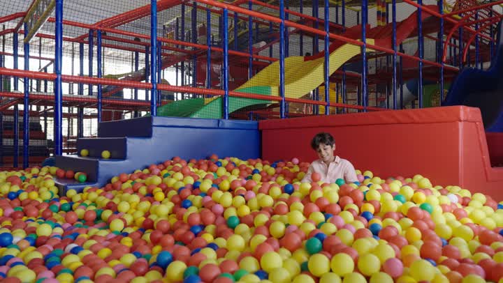 Sitting happily in the small ball pit, a young Arab Gulf Saudi boy is digging a hole among the plastic balls, with shots filled with joy and fun, enjoying playing in the amusement park.