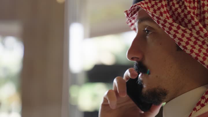 النظرات الجدية اثناء اجراء مكالمة هاتفية , رجل عربي سعودي خليجي يرتدي الزي التقليدي يتحدث في الهاتف المحمول ,استخدام التقنيات الحديثة للبقاء متصلين مع العالم الخارجي ,تصوير جانبي مقرب