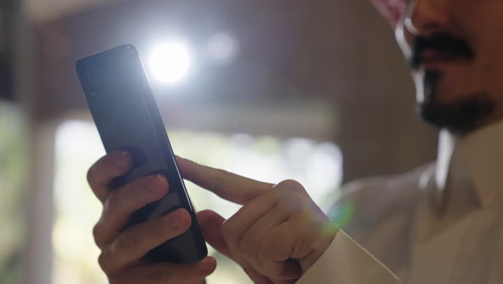 Spending enjoyable times with entertainment games, an Arab Gulf Saudi man wearing traditional attire holding a mobile phone with gestures of happiness indicating a good time, using modern technology in enjoyable fields in addition to communications and knowing the news of the outside world.