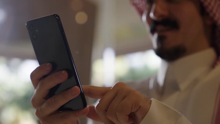 استخدام التقنية الحديثة  في معظم جوانب الحياة اليومية ,شاب عربي سعودي خليجيى يرتدي الزي التقليدي السعودي يتصفح التطبيقات المسلية المختلفة في الهاتف المحمول , ايماءات السعادة و البهجة اشارة الى قضاء اوقات ممتعة