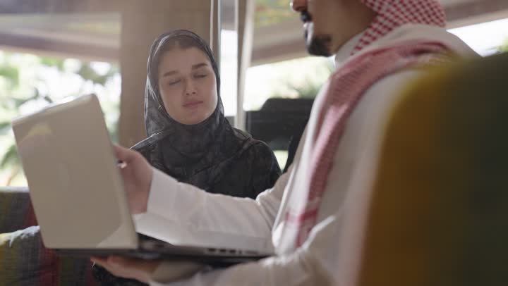 شاب عربي سعودي خليجي يرتدي الزي السعودي التقليدي يشرح لاخته طريقة التعامل مع الحاسب ,فتاة عربية خليجية سعودية تستمع بانصات لاخيها ,اكتساب مهارات جديدة ,التعلم و زيادة المعرفة بالاجهزة المتطورة