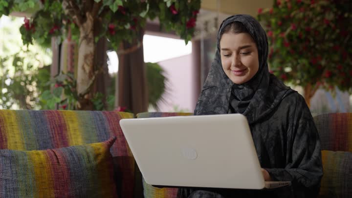 Following family news and communicating with the outside world from home, using modern and advanced electronic devices easily, an Arab Gulf Saudi girl wearing a gray abaya sits on the living room sofa, communicating with friends through social media.