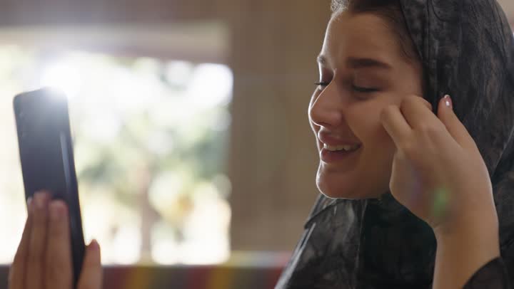 التواصل بسهولة مع الاصدقاء و العائلة باستخدام تقنيات التكنولوجيا الحديثة , تطبيقات الهاتف المحمول المتطورة تساعد في اجراء مكالمات الفيديو و البقاء متصلين للاطمئنان على العائلة ,فتاة عربية خليجية سعودية محجبة ترتدي عباءة رمادية تجري اتصال بخاصية الفيديو بسعادة و فرحة