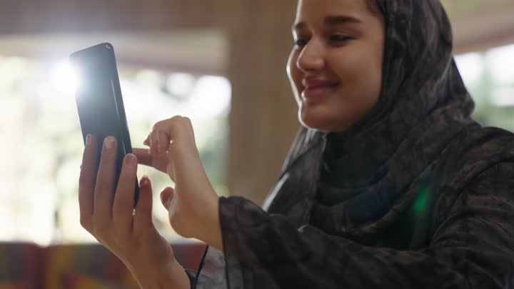 قضاء اوقات مسلية اثناء التصفح على الهاتف المحمول ,فتاة عربية خليجية سعودية محجبة ترتدي عباءة رمادية تقوم باستخدام مواقع التواصل الاجتماعي و تطبيقات الالعاب المسلية ,البقاء متصلين مع العائلة و الاصدقاء بسهولة