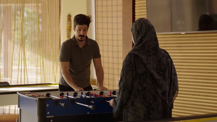 Engaging in a collective recreational activity for the whole family, a young Saudi Arabian Gulf Arab is skillfully maneuvering the handles of a foosball game to score goals, with looks of determination and seriousness while maintaining a connected family atmosphere, on a beautiful sunny day, enjoyable family entertainment activities.