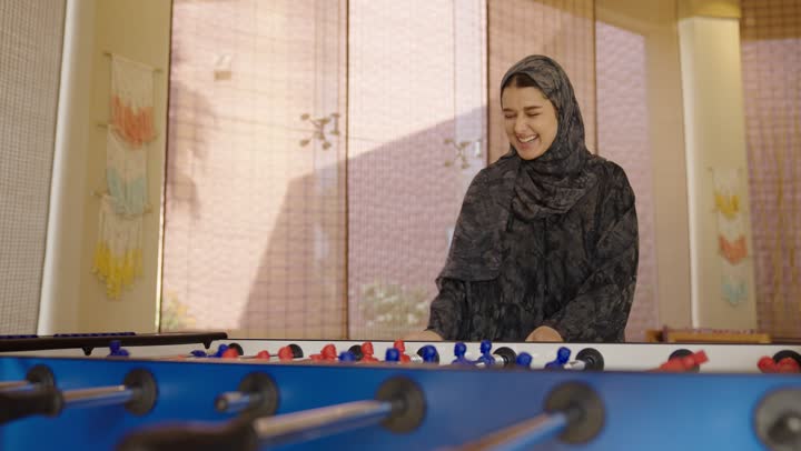 اجراء حديث شيق ممتع اثناء لعب الفوسبول ,لحظات مليئة بايماءات السعادة و البهجة الواضحة ,فتاة عربية خليجية سعودية محجبة ترتدي عباءة رمادية تتحكم بالمقابض باحترافية عالية لتسجيل الاهداف