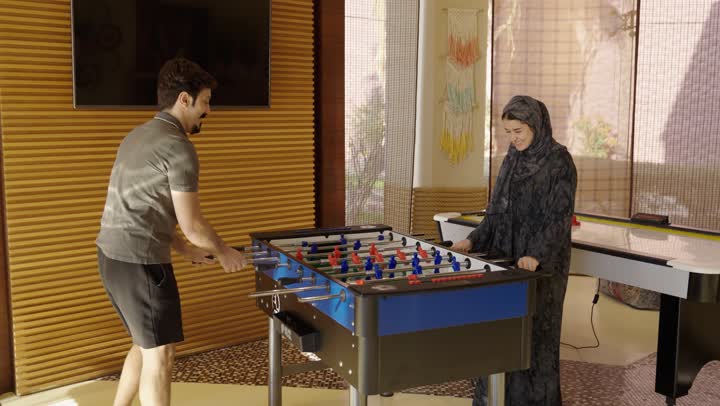 قضاء اوقات عائلية مرحة اثناء لعب الفوسبول ,شاب عربي خليجي سعودي يرتدي الزي الكاجوال يحرك مقابض التحكم بسرعة لتحقيق الفوز ,فتاة عربية خليجية سعودية محجبة ترتدي عباءة رمادية تقوم بصد الهجمات و تحقيق انتصار علي اخيها ,جو اسري ملئ بالضحك و السعادة في يوم مشمس دافئ