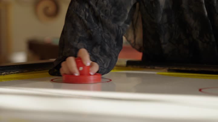 الامساك بالمضرب الخاص بلعبة الهوكي الهوائي في حركات تبادلية بين اللاعبين لاتمام اللعب و تحقيق الفوز ,فتاة عربية خليجية سعودية ترتدي عباءة رمادية تقوم باللعب مع الوالد في جو اسري ممتع