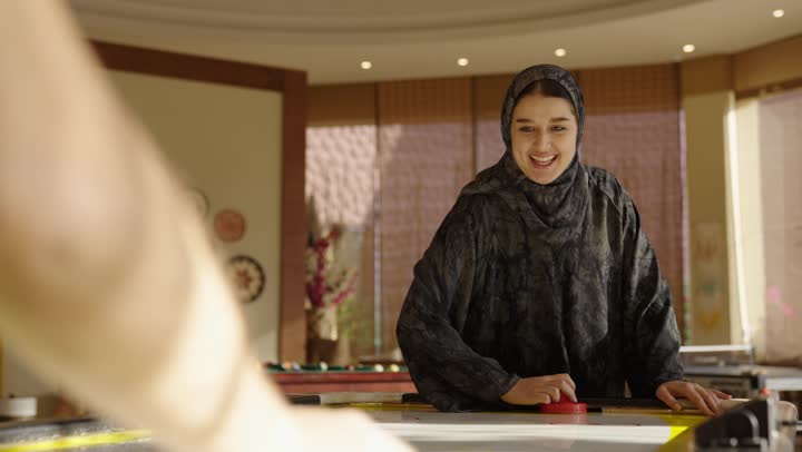 A Saudi Arabian Gulf girl wearing a gray abaya is having fun playing air hockey, competing with her father to create unforgettable family moments, cheering and clapping after achieving victory.