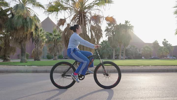 فتاة عربية خليجية سعودية ترتدي الزي الكاجوال تقود الدراجة الهوائية ,التنزه في الحدائق الخضراء ,الاستمتاع بالجو الدافئ والسماء الصافية
