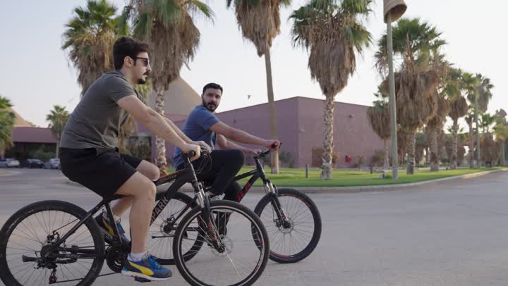 حديث عائلي شيق بين الوالد والابن اثناء ركوب الدراجة الهوائية ,مفهوم الترابط الاسري ,رجل عربي سعودي خليجي يرتدي الزي الكاجوال ,شاب يرتدي السروال القصير و النظارة الشمسية في جولة بالدراجات وسط الاشجار و النخيل ,الطبيعة الساحرة