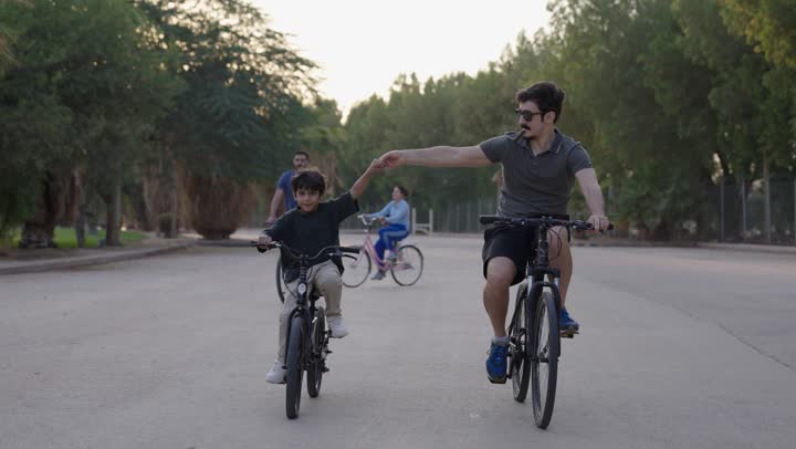 Enjoying the pleasant and moderate atmosphere amidst nature and trees, an Arab Saudi Gulf man dressed in casual attire rides a bicycle with his young son, a small child holding his father's hand to feel safe while riding the bike, a girl skillfully riding the wheel with her siblings.