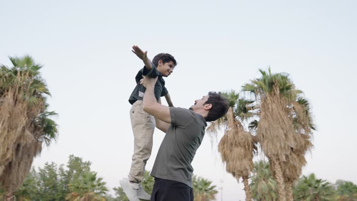 The concept of safety and reassurance, an Arab Saudi Gulf man wearing casual attire holds his son in the air happily and carefully, a small son smiling at his father feeling love, spreading his arms and flying in the sky, the enchanting nature and clear sky.