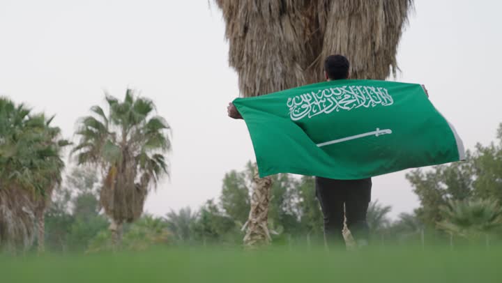 راية التوحيد الخضراء ترفرف في الهواء عالياً ,رجل عربي سعودي خليجي يرتدي الزي الكاجوال يحمل علم المملكة العربية السعودية ,الاستمتاع بالاشجار و الطبيعة