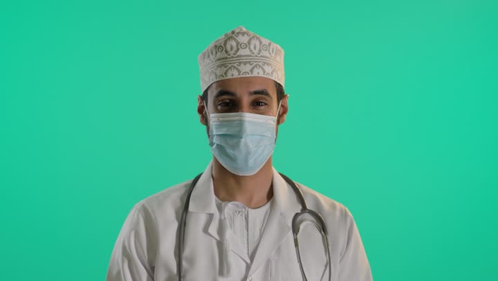 السلامة والحماية من فايروس كورونا، اتباع اجراءات الوقاية من كوفيد19, بورتريه لطبيب عربي خليجي عماني يرتدي الدشداشة و الكمة و المعطف الطبي والقفازات والكمامة لضبط انتشار العدوى وسماعة طبية يقف بايدي متقاطعة , خلفية خضراء ، كروما