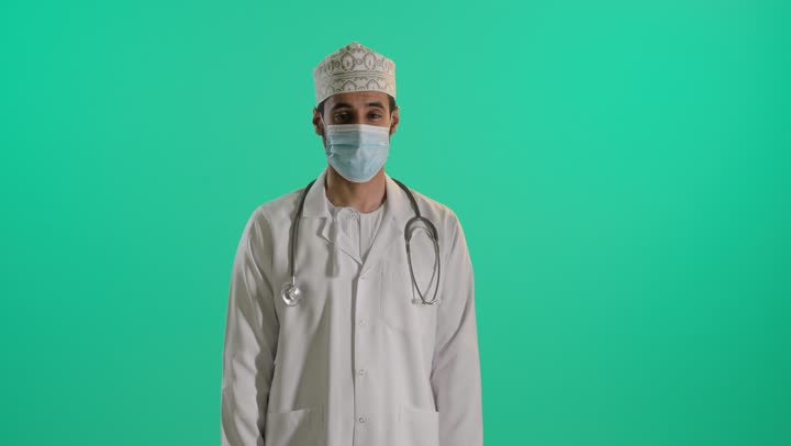 اتباع اجراءات الوقاية من كوفيد19, السلامة والحماية من فايروس كورونا, بورتريه لطبيب عربي خليجي عماني يرتدي الدشداشة و الكمة و المعطف الطبي والقفازات والكمامة لضبط انتشار العدوى وسماعة طبية يقف بايدي متقاطعة , خلفية خضراء ، كروما