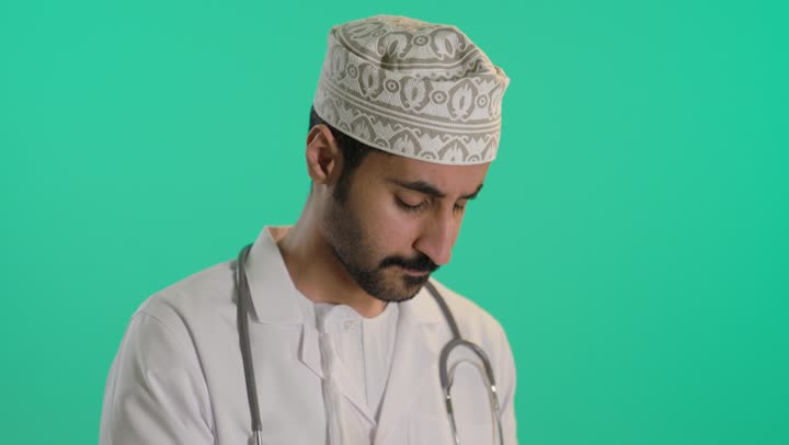 Thinking and trying to remember something, working in centers and hospitals, medicine and healthcare, taking notes and data, a portrait of an Arab Gulf Omani doctor wearing a dishdasha and kumma and a lab coat, putting on a stethoscope and writing, green background, chroma.
