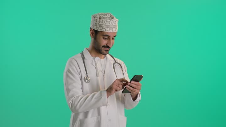 Looking at the camera with a joyful expression, using modern technology in the medical field, providing therapeutic services, a portrait of an Arab Gulf Omani doctor wearing a dishdasha and kumma and a medical coat, wearing a stethoscope and using a mobile phone, green background, chroma.