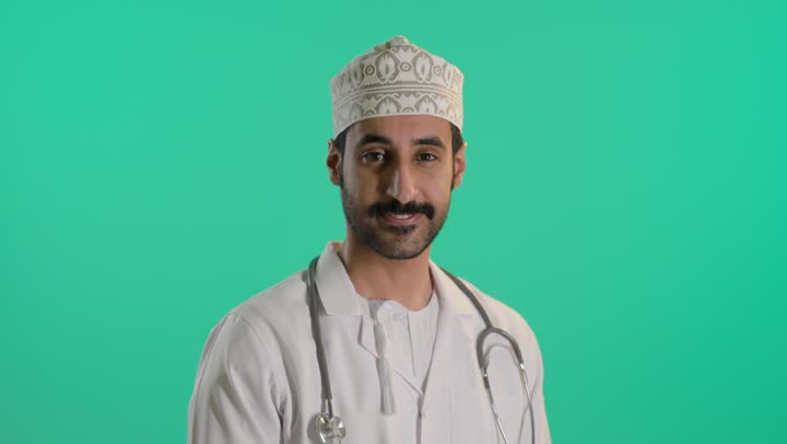 Looking with gestures of self-confidence, medical examination and providing therapeutic services to patients, a portrait of an Arab Gulf Omani doctor wearing a dishdasha and kumma and a medical coat, holding a stethoscope, standing with arms crossed, green background, chroma.