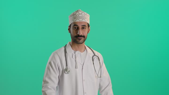 Medical examination and providing treatment services to patients, a portrait of an Arab Gulf Omani doctor wearing a dishdasha and kumma and a medical coat, holding a stethoscope, standing with arms crossed, looking with gestures of self-confidence, green background, chroma.