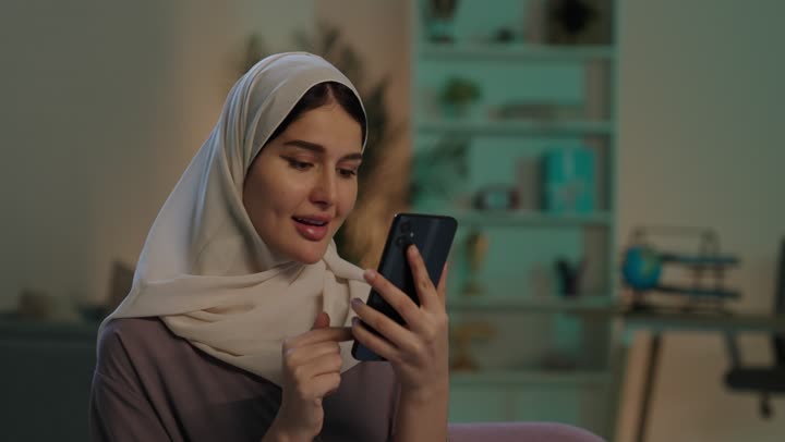 الدردشه مع الأهل والأصدقاء، إيماءات السرور والسعادة، تصفح تطبيقات وبرامج التواصل الاجتماعي، تصوير مقرب لشابة عربية خليجية سعودية محجبة مبتسمة ترتدي العباءة و الحجاب تمسك بيدها الهاتف المحمول، استخدام الاجهزة التقنية الحديثة والمتطورة