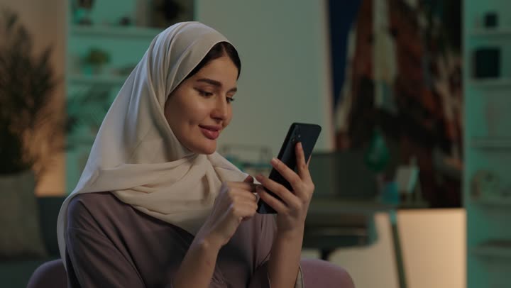 إيماءات السرور والسعادة، تصفح تطبيقات وبرامج التواصل الاجتماعي، الدردشه مع الأهل والأصدقاء، تصوير مقرب لشابة عربية خليجية سعودية محجبة مبتسمة ترتدي العباءة و الحجاب تمسك بيدها الهاتف المحمول، استخدام الاجهزة التقنية الحديثة والمتطورة