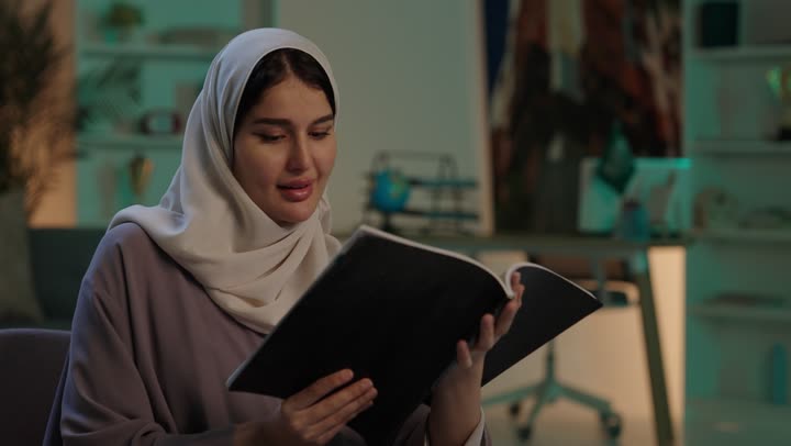 Spending free time in a useful way, developing skills and expanding knowledge and ideas, enjoying reading books, stories, and novels, a young Arab Gulf Saudi woman wearing an abaya and hijab holding a book in her hands, increasing intellectual resources.