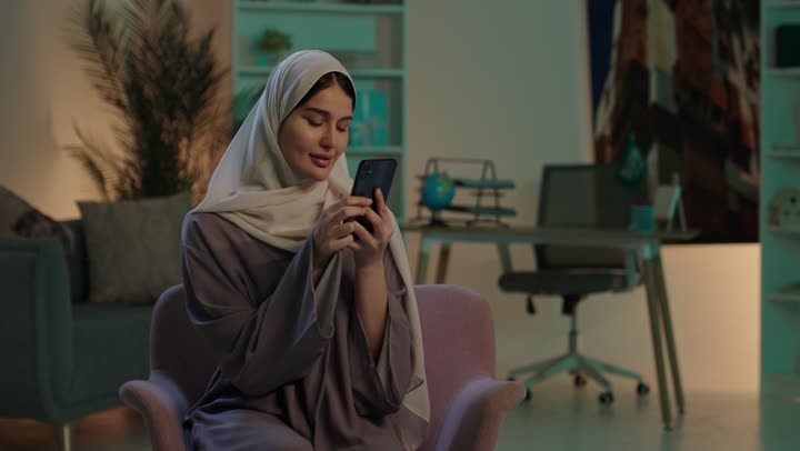 النظر الى الكاميرا بتعابير السرور، التسوق الالكتروني عبر الانترنت، استخدام البرامج والتطبيقات الالكترونية، تصفح المواقع الالكترونية والتواصل الاجتماعي،  شابة عربيه خليجيه سعودية ترتدي الحجاب و العباءة تجلس على كرسي تستخدم الهاتف المحمول