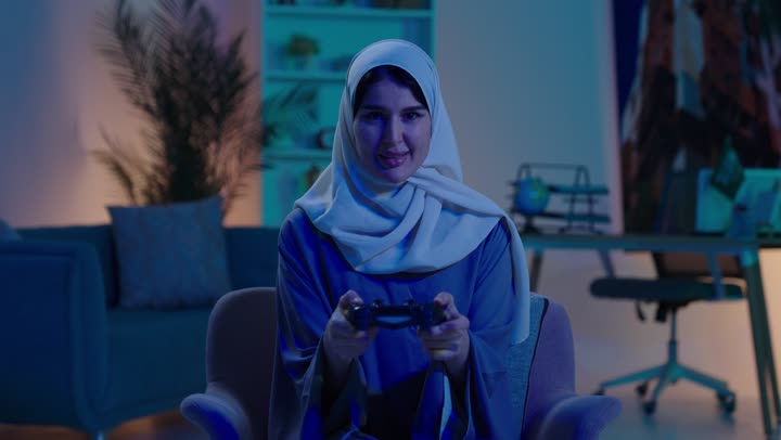 قضاء وقت ممتع ومسلي، التفاعل والاندماج مع ألعاب الفيديو ،اجهزة وتقنيات الكترونية حديثة ترفيهيه, ايماءات الفوز و السعادة ، شابة عربية خليجية سعودية ترتدي العباءة و الحجاب تجلس  على كرسي تحمل عصا التحكم