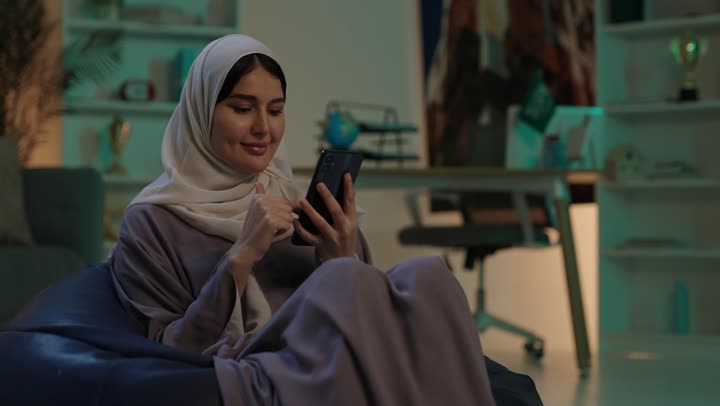 التسوق الالكتروني عبر الانترنت، استخدام البرامج والتطبيقات الالكترونية، تصفح المواقع الالكترونية والتواصل الاجتماعي،  شابة عربيه خليجيه سعودية ترتدي الحجاب و العباءة تجلس على كرسي بين باج تستخدم الهاتف المحمول