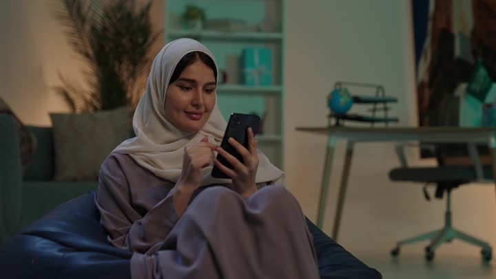 Expressions of happiness and joy, online shopping, using electronic programs and applications, browsing websites and social media, a young Arab Gulf Saudi woman wearing a hijab and abaya sitting on a chair between bags using a mobile phone.