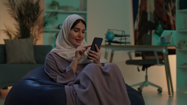 استخدام البرامج والتطبيقات الالكترونية، تصفح المواقع الالكترونية والتواصل الاجتماعي،  شابة عربيه خليجيه سعودية ترتدي الحجاب و العباءة تجلس على كرسي بين باج تستخدم الهاتف المحمول ، التسوق الالكتروني عبر الانترنت