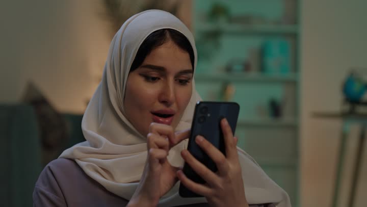 تصفح تطبيقات وبرامج التواصل الاجتماعي، الدردشه مع الأهل والأصدقاء، إيماءات السرور والسعادة، تصوير مقرب لشابة عربية خليجية سعودية محجبة مبتسمة ترتدي العباءة و الحجاب تمسك بيدها الهاتف المحمول، استخدام الاجهزة التقنية الحديثة والمتطورة
