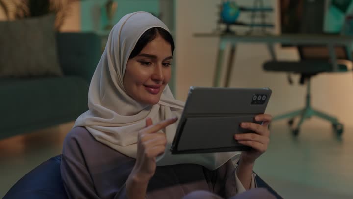Chatting with family and friends, integrating modern technologies and devices into practical life, a young Arab Gulf Saudi woman wearing an abaya and hijab sits on a chair with a bag, holding a tablet in her hands, browsing websites.