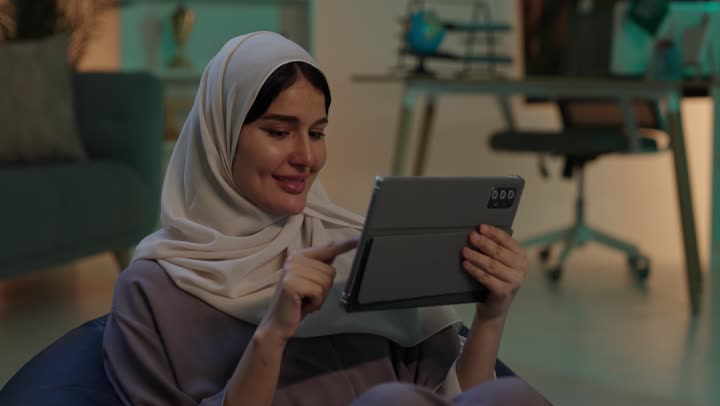 النظر الى الكاميرا بتعابير السرور، الدردشة مع الأهل والأصحاب، دمج التقنيات الحديثة والأجهزة التكنولوجية بالحياة العملية، شابة عربية خليجية سعودية ترتدي العباءة و الحجاب تجلس على كرسي بين باج تمسك  جهاز التابلت بيديها، تصفح مواقع الانترنت