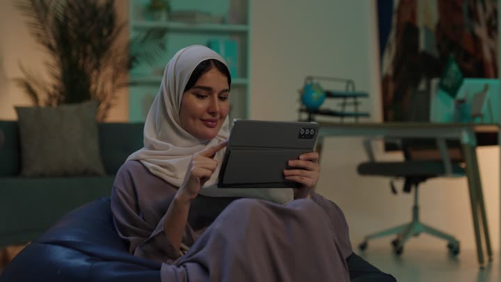 دمج التقنيات الحديثة والأجهزة التكنولوجية بالحياة العملية، شابة عربية خليجية سعودية ترتدي العباءة و الحجاب تجلس على كرسي بين باج تمسك  جهاز التابلت بيديها، تصفح مواقع الانترنت، الدردشة مع الأهل والأصحاب
