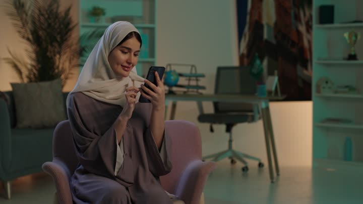 الدردشه مع الأهل والأصدقاء، تصفح تطبيقات وبرامج التواصل الاجتماعي، إيماءات السرور والسعادة، شابة عربية خليجية سعودية محجبة مبتسمة ترتدي العباءة و الحجاب تجلس على كرسي تمسك بيدها الهاتف المحمول، استخدام الاجهزة التقنية الحديثة والمتطورة