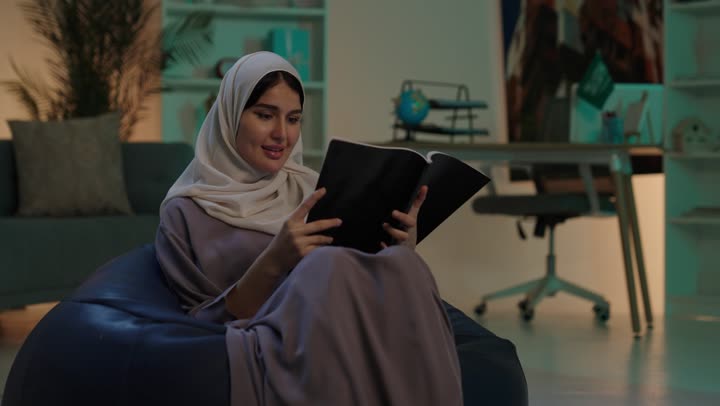 مفهوم الشغف بالمطالعة، الاستمتاع بقراءة الكتب والقصص والروايات، قضاء الوقت بالتعلم والتثقف، شابة عربية خليجية سعودية ترتدي العباءة و الحجاب تجلس على كرسي بين باج تحمل بيديها كتاب، ثقافة التعلم الذاتي