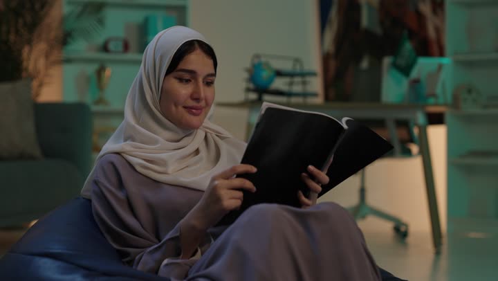 الاستمتاع بقراءة الكتب والقصص والروايات, مفهوم الشغف بالمطالعة، قضاء الوقت بالتعلم والتثقف، شابة عربية خليجية سعودية ترتدي العباءة و الحجاب تجلس على كرسي بين باج تحمل بيديها كتاب، ثقافة التعلم الذاتي