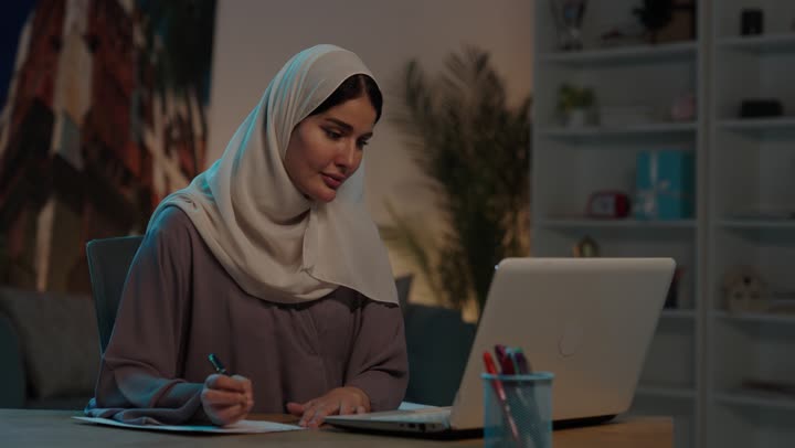 Using white paper to record notes and important information, typing on the keyboard, the concept of writing and taking notes, a young Arab Gulf Saudi woman wearing an abaya and hijab holding a pen writing on a notebook and working on a laptop.