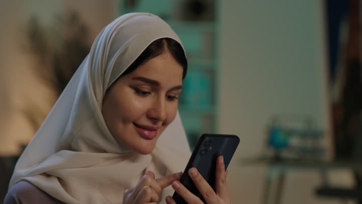 Following social media platforms, a close-up shot of a Saudi Arabian Gulf woman wearing a hijab and an abaya, holding a mobile phone in her hands, using modern technology, with gestures of happiness and joy.