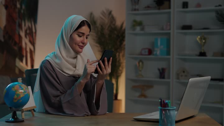 استخدام الأجهزة والتقنيات الحديثة المتطورة، تصفح التطبيقات وبرامج التواصل الاجتماعي، تبادل الأحاديث والأخبار مع الأهل والأصدقاء، شابة عربية خليجية سعودية محجبة ترتدي العباءة تجلس امام الحاسب المحمول  تمسك بيدها الجوال