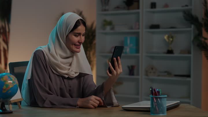 اجراء مكالمة فيديو من خلال الهاتف الجوال، التقاط صور سيلفي, التواصل والتحدث مع الأهل والأصدقاء, شابة عربية خليجية سعودية ترتدي العباءة والحجاب تمسك بيدها الهاتف المحمول، ايماءات السعادة و السرور