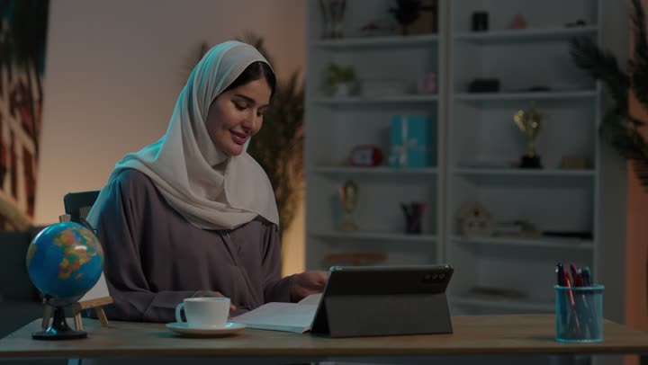 الاستمتاع بتناول المشروب المفضل، مراجعة الدروس اليومية عبر الانترنت، تطور العلم والتعليم في المملكة السعودية،  شابة عربية خليجية سعودية محجبة ترتدي العباءة تجلس امام جهاز التابلت المحمول تقوم بقراءة كتاب، مذاكرة الدروس وتلقي المحاضرات عن بعد