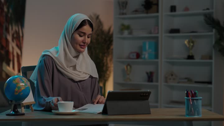 مراجعة الدروس اليومية عبر الانترنت، تطور العلم والتعليم في المملكة السعودية،  شابة عربية خليجية سعودية محجبة ترتدي العباءة تجلس امام جهاز التابلت المحمول تقوم بقراءة كتاب، مذاكرة الدروس وتلقي المحاضرات عن بعد، الاستمتاع بتناول المشروب المفضل ، ايماءات السعادة و السرور